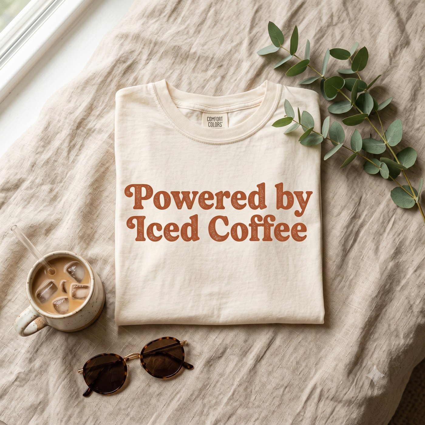 A real Etsy-listing-style styled flat-lay: a folded comfort-colors tee propped with eucalyptus, a mug and sunglasses. Any styled mockup works; a bare shirt on white does not.